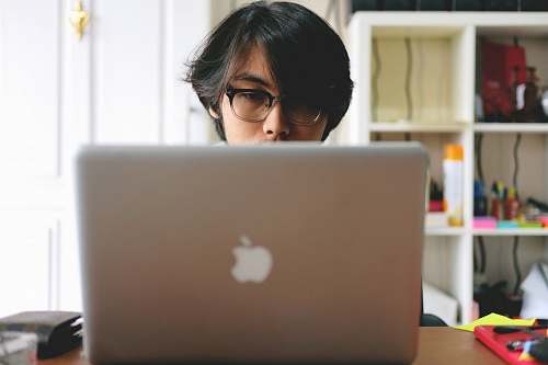 person man wearing eyeglasses while using silver Macbook near wooden cubby shelf inside room people