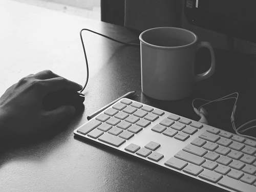 black-and-white grayscale photography of person holding computer mouse near keyboard and mug grey