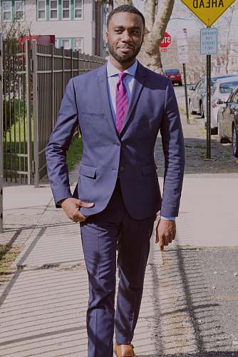person man in blue suit walking on the sidewalk near metal fence and parked vehicle human