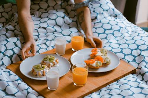 meal person serving pastries on white ceramic plates with fruit juice glasses on wooden tray on top of bed breakfast