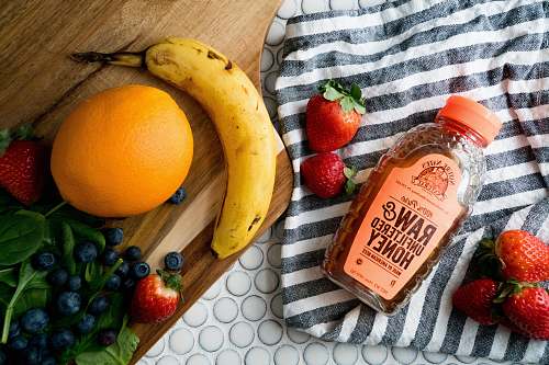 fruit raw and unfiltered honey bottle between strawberries with blueberries, citrus, and banana fruits nearby blueberry