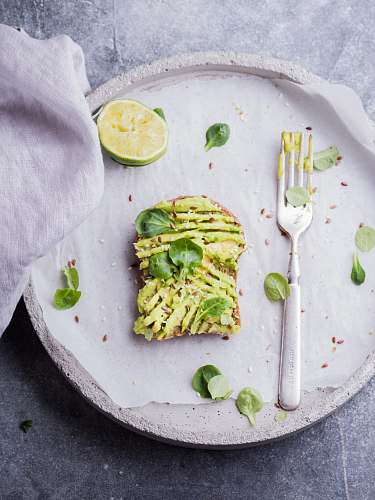 plate wheat bread with avocado spread beside white plastic spoon and pressed lime on round white plate breakfast