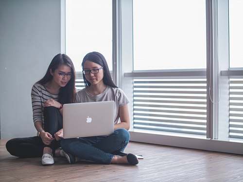 person two women facing silver MacBook sitting on parquet floor people