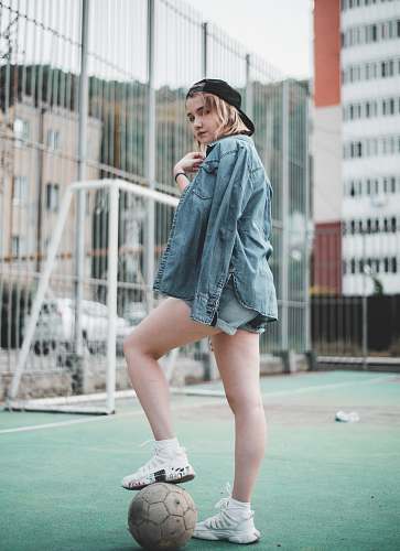 apparel woman wearing blue denim jacket standing and stepping right foot on white soccer ball in green field during daytime clothing