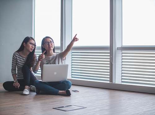 laptop two women sitting on brown floor inside white room beside glass windows pointing upwards photo computer