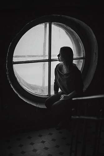 window grayscale photo of woman sits near round wooden framed window person