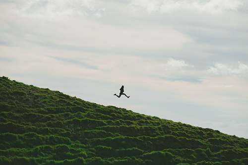 hill person jumping on grass field under cloudy sky during daytime jumping