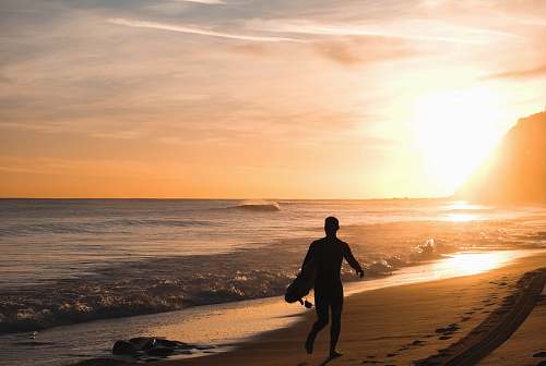 person silhouette of man carrying surfboard running on shore people