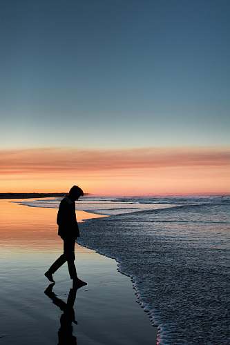 person silhouette of man walking on seashore under orange sky standing