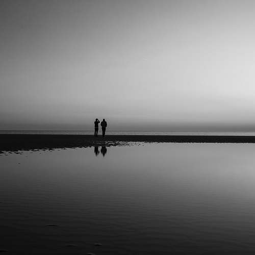 person silhouette of two person standing beside calm water black-and-white