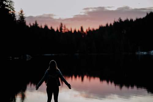 person woman standing with hands spread on side facing lake at sundown silhouette