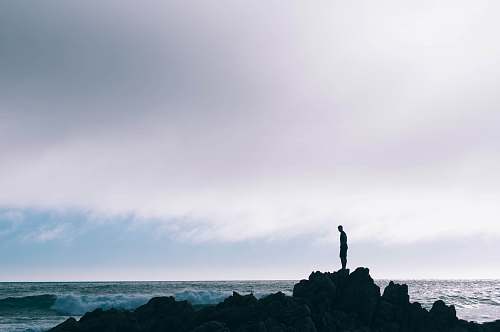 silhouette man standing on boulder in front of ocean standing