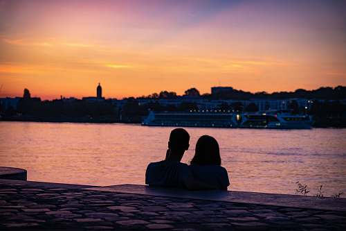 human silhouette of couple sitting on shore looking at city skyline during sunset sitting