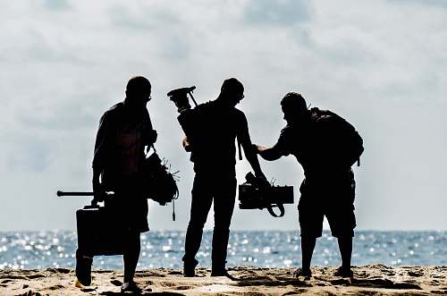 silhouette silhouette of men holding camera standing on sand near body of water during daytime human
