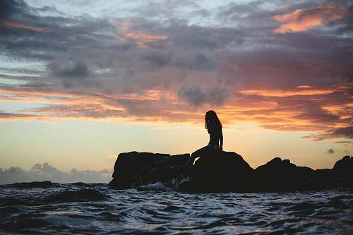 woman silhouette photo of woman sitting on rock during golden hour water