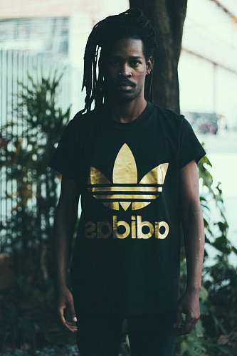 person depth of field photography of man wearing black and gold adidas graphic crew-neck T-shirt standing in front of tree people