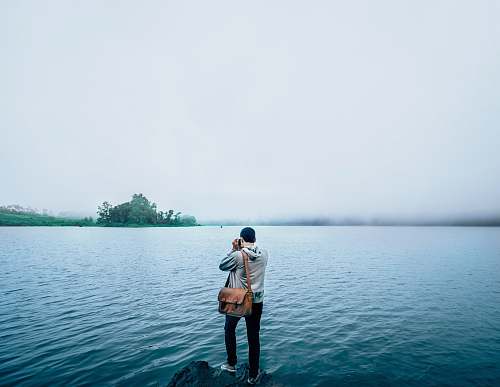 person man holding brown leather saddlebag standing on edge of rock over body of water people