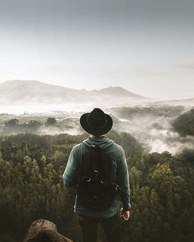person man standing on mountain top overlooking green-leafed trees at foggy daytime people