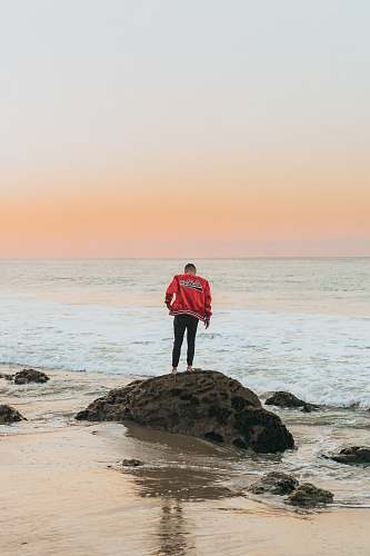 person man wearing orange jacket standing on seashore rock near beach people