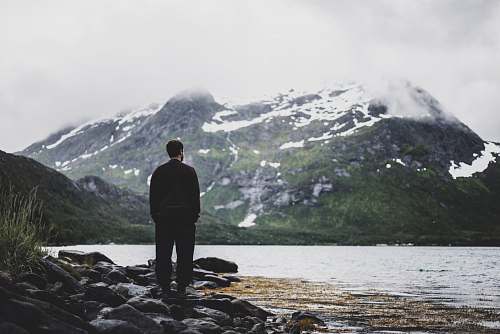 people photo of man standing near seashore facing mountain covered with snow person