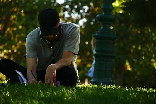 people selective focus photography of man sitting on grass and writing on notebook during daytime person
