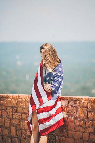 people woman standing leaning on brick wall wrapping body with U.S. flag near body of water person