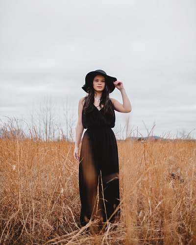 people woman wearing black V-neck spaghetti-strap jumpsuit and black hat standing on brown grass field person