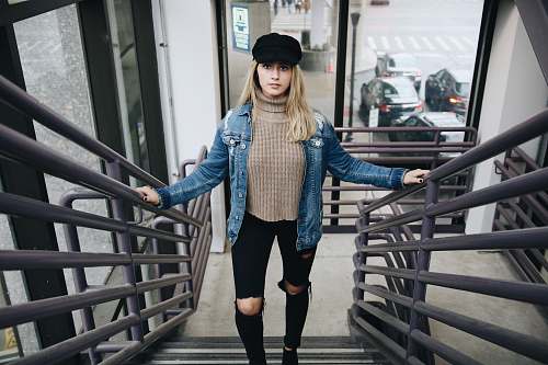 people woman wearing blue denim jacket, brown sweater, black jeans, and hat standing in stair person
