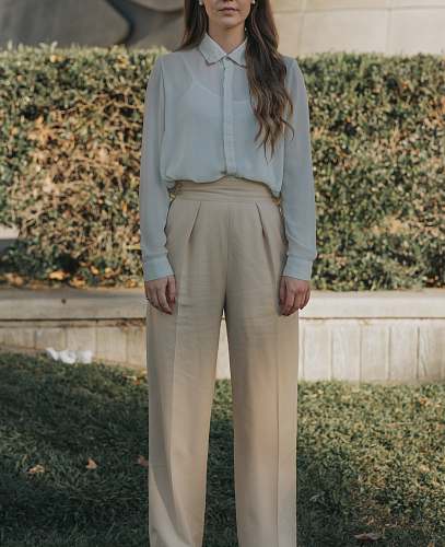 people woman wearing gray button-up long-sleeved shirt and brown pants standing in front of bushes person