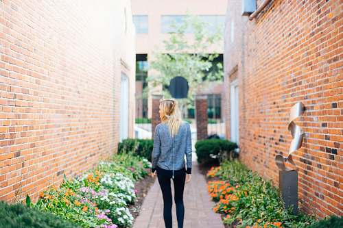 person woman wearing gray long-sleeved shirt and black fitted pants in between of brown bricked walls with flowers during daytime people