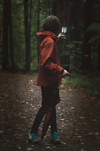 person woman wearing red hoodie holding smartphone near green leafed tree people