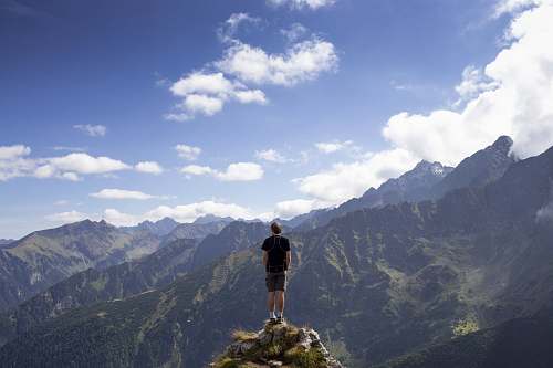 mountain man wearing black shirt and gray shorts on mountain hill beside mountains under white and blue cloudy skies mountain range