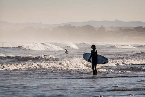 sea person holding white surfboard standing on body of water under gray sky during daytime surfboard