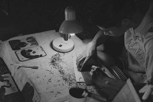 black-and-white grayscale photography of man writing on brown paper near turned on lamp grey