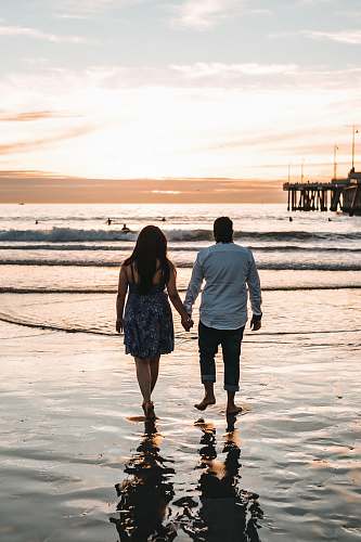 human man and woman holding hands each other while walking on seashore during daytime pier
