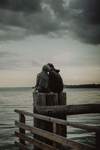 human man and woman sitting and kissing on post near the body of water person