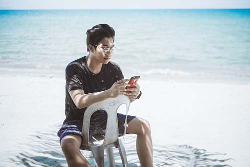 human man in black shirt and blue board shorts sitting on plastic chair near seashore person