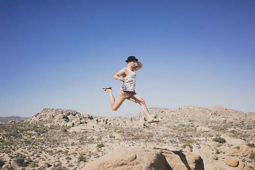 person man jumping on brown stone while holding his hat desert