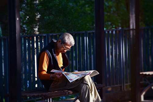 human man reading newspaper while sitting on bench in front of wooden fence person