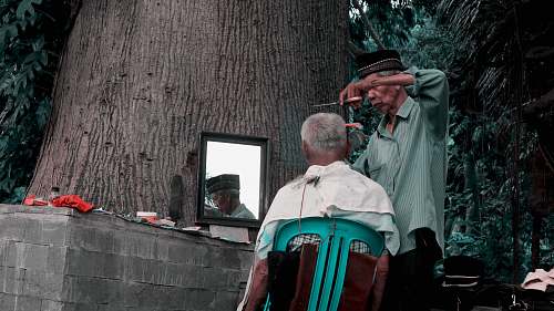 human man sitting on green chair facing mirror while the other man standing cutting's man hair at daytime person