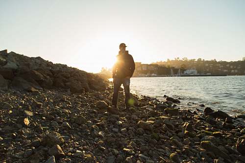 sun man wearing black jacket standing on stones near body of water under blue sky during golden hour rock