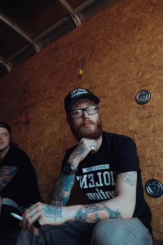 human man with cigarette stick on the hand while holding beard sitting near wall tattoo