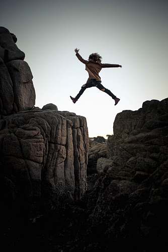 person person jumping on big rock under gray and white sky during daytime rock