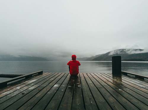 human person wearing red hooded jacket sitting on brown and black wooden sea dock near mountains under gray sky during daytime person