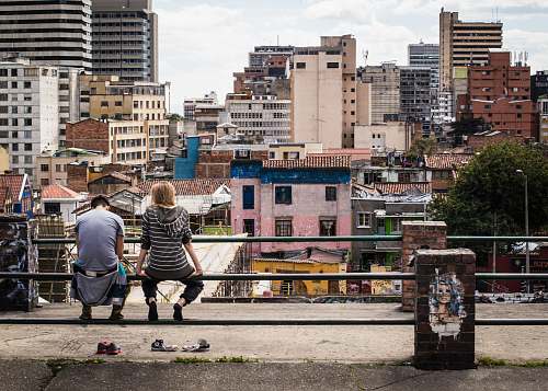 human photo of two people sitting on pole fence facing high-rise building person