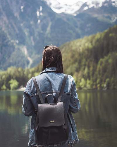 human selective focus photography of woman wearing blue denim jacket and black bucket backpack near lagoon at daytime person