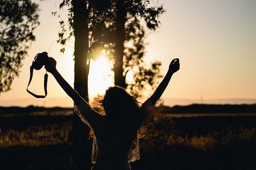 human silhouette photo of woman raising her hands while holding DSLR camera person