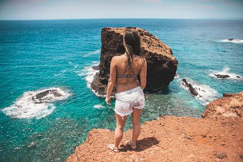 human woman in white denim shorts standing on rock facing sea person