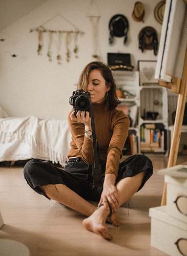 person woman sitting on brown wooden floor while holding black DSLR camera in room human