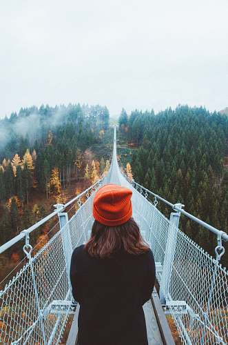 human woman wearing knit cap walking on white bridge between trees during daytime bridge